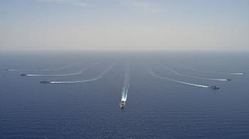 Seven naval ships sailing in formation in open water with the horizon behind them,