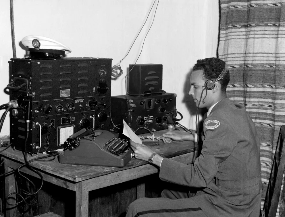 Black and white photograph of a young man in a military peacekeeping uniform siting at a desk with a headset on and a typewriter and other communication equipment in front of them.