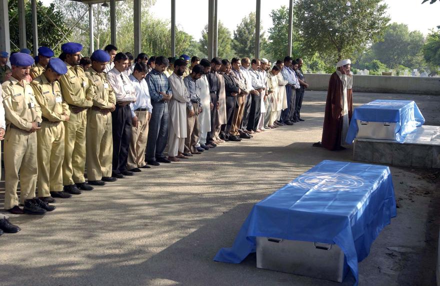 People stand in rows for a prayer near two coffins covered with UN flags, under a shaded structure.