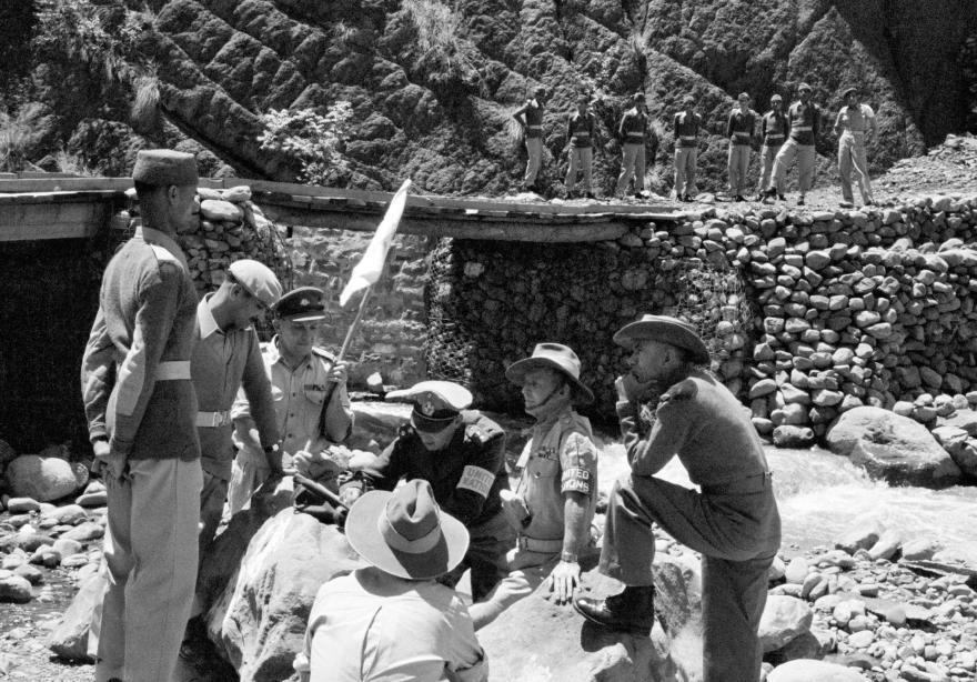 UN military observers and local soldiers gather outdoors near a rocky stream and a stone bridge. Some stand while others sit on large rocks. A white flag is visible among the group, and additional soldiers are lined up on the bridge above.
