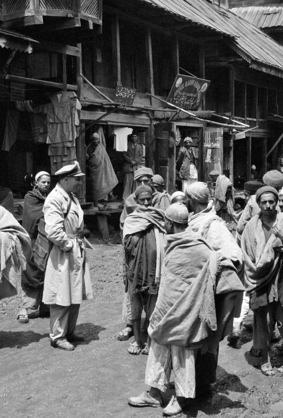 UN military observer in uniform speaks with a group of people wrapped in blankets in a rural outdoor setting with wooden structures in the background.
