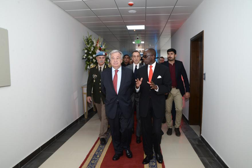 UN Secretary General Guterres walking down a hallway with Chief of Mission Support (CMS) Mr. Nester Odaga-Jalomayo. Behind them are a group of people following them.