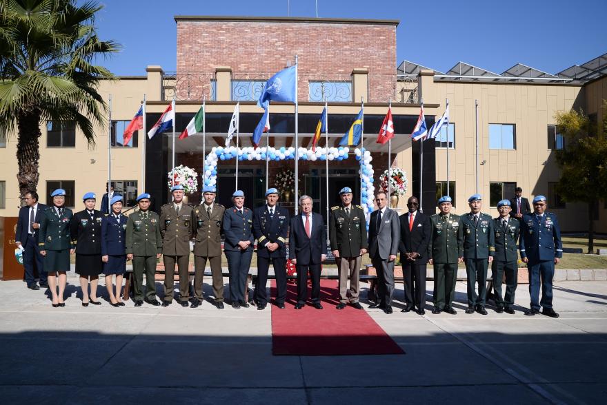UN military personnel in uniform and blue berets stand in a line on a red carpet outside a building with UN and national flags