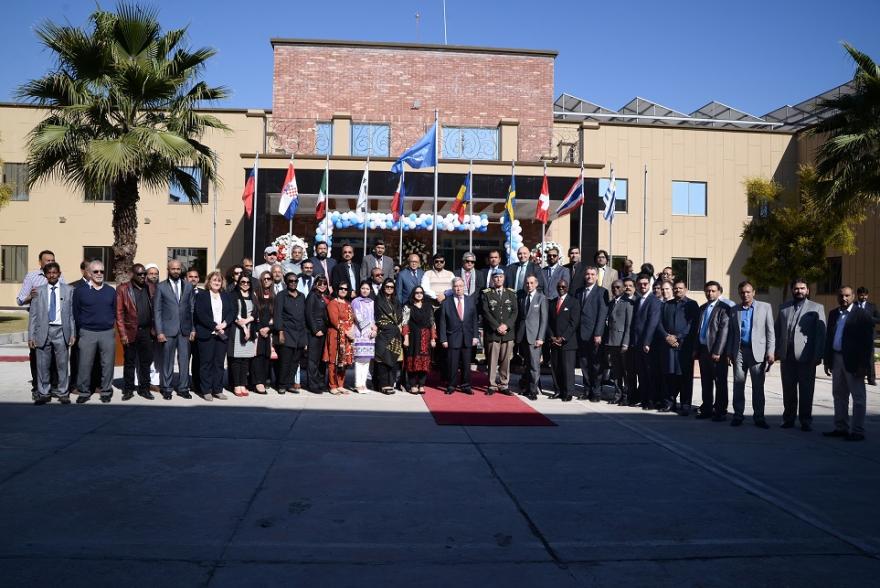 UN military personnel in uniform and blue berets stand in a line on a red carpet outside a building with UN and national flags