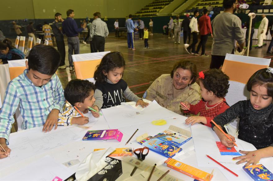 A woman crouched down with some children seated at a table indoors drawing and coloring on large sheets of paper with crayons and pencils during an art activity.