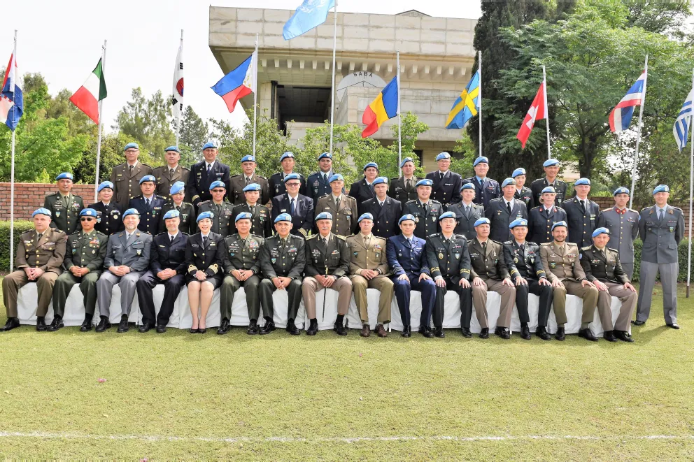 Group photo of UN military personnel in uniform and blue berets seated and standing in rows outdoors with national flags behind them.