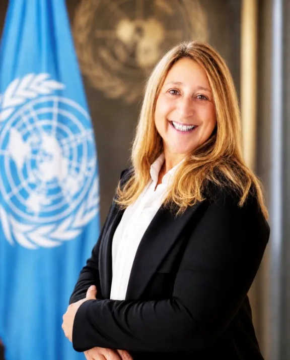 Portrait of a woman smiling, wearing a white collared blouse and black suit jacket. In the background is the UN flag and UN emblem.